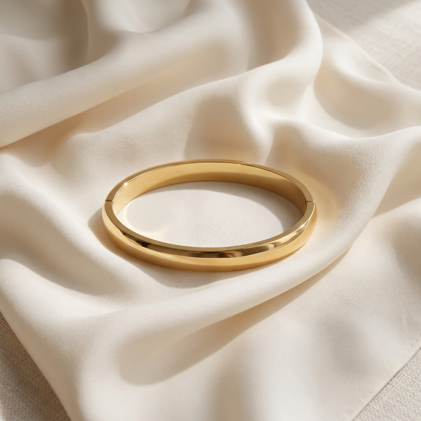 A polished gold bangle bracelet resting on draped cream-colored satin fabric.