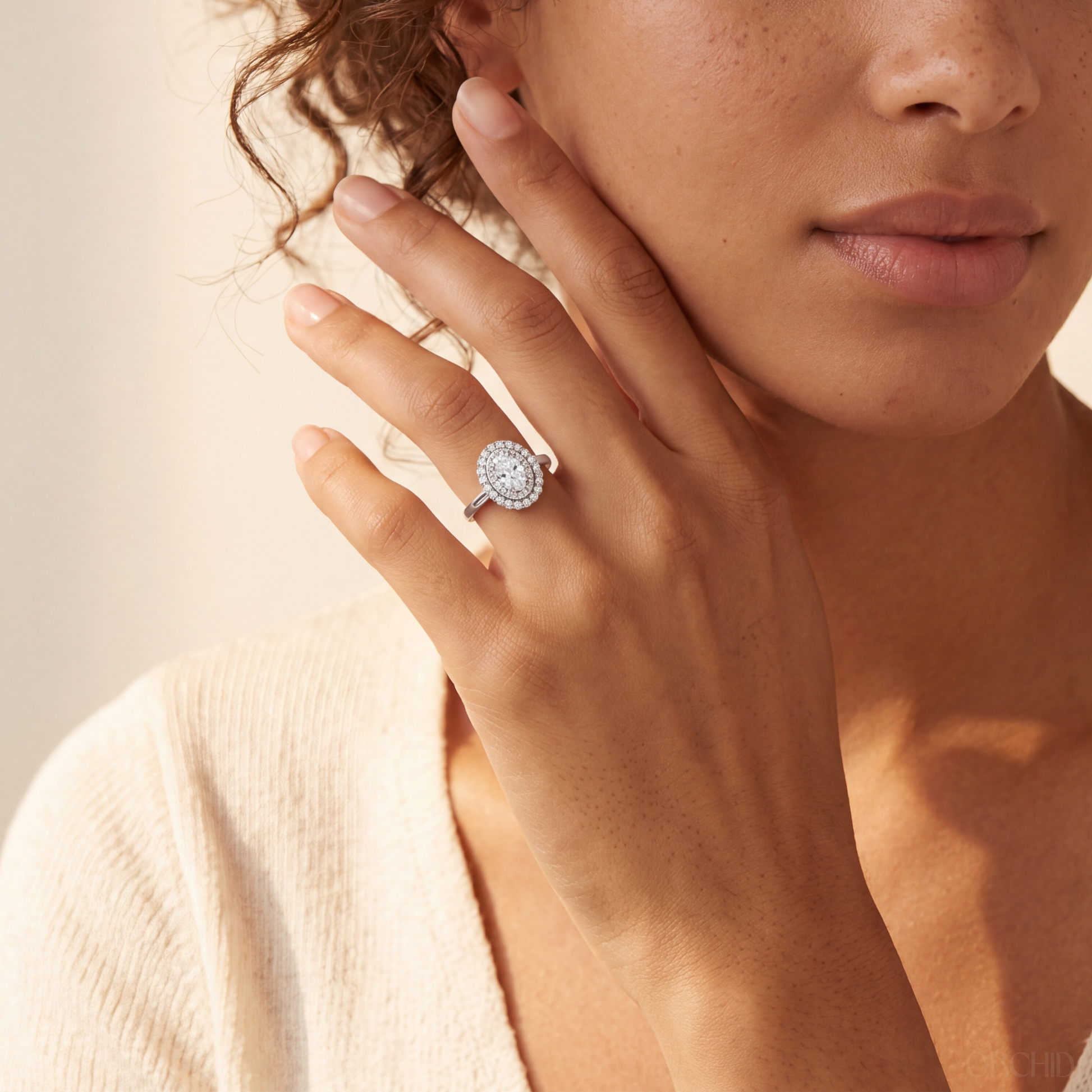 A close-up lifestyle shot of a woman wearing a silver halo ring with a round-cut diamond center stone on her finger, set against a warm beige background.