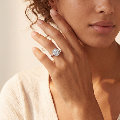 A close-up lifestyle shot of a woman wearing a silver halo ring with a round-cut diamond center stone on her finger, set against a warm beige background.