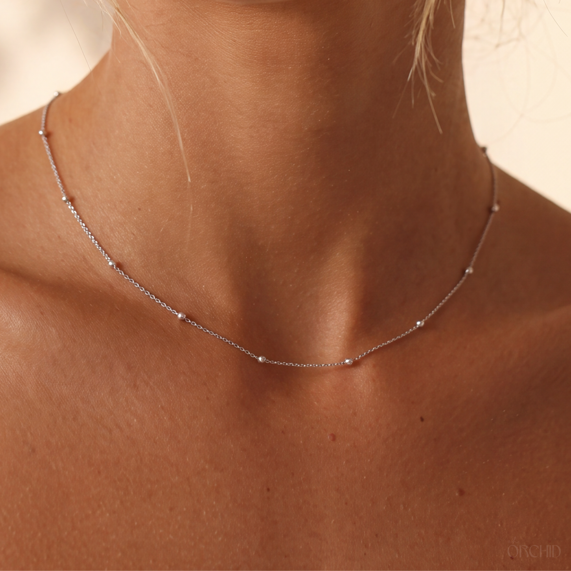 A delicate silver satellite beaded chain necklace worn on a model's neck, shown in a close-up shot with warm, soft lighting.