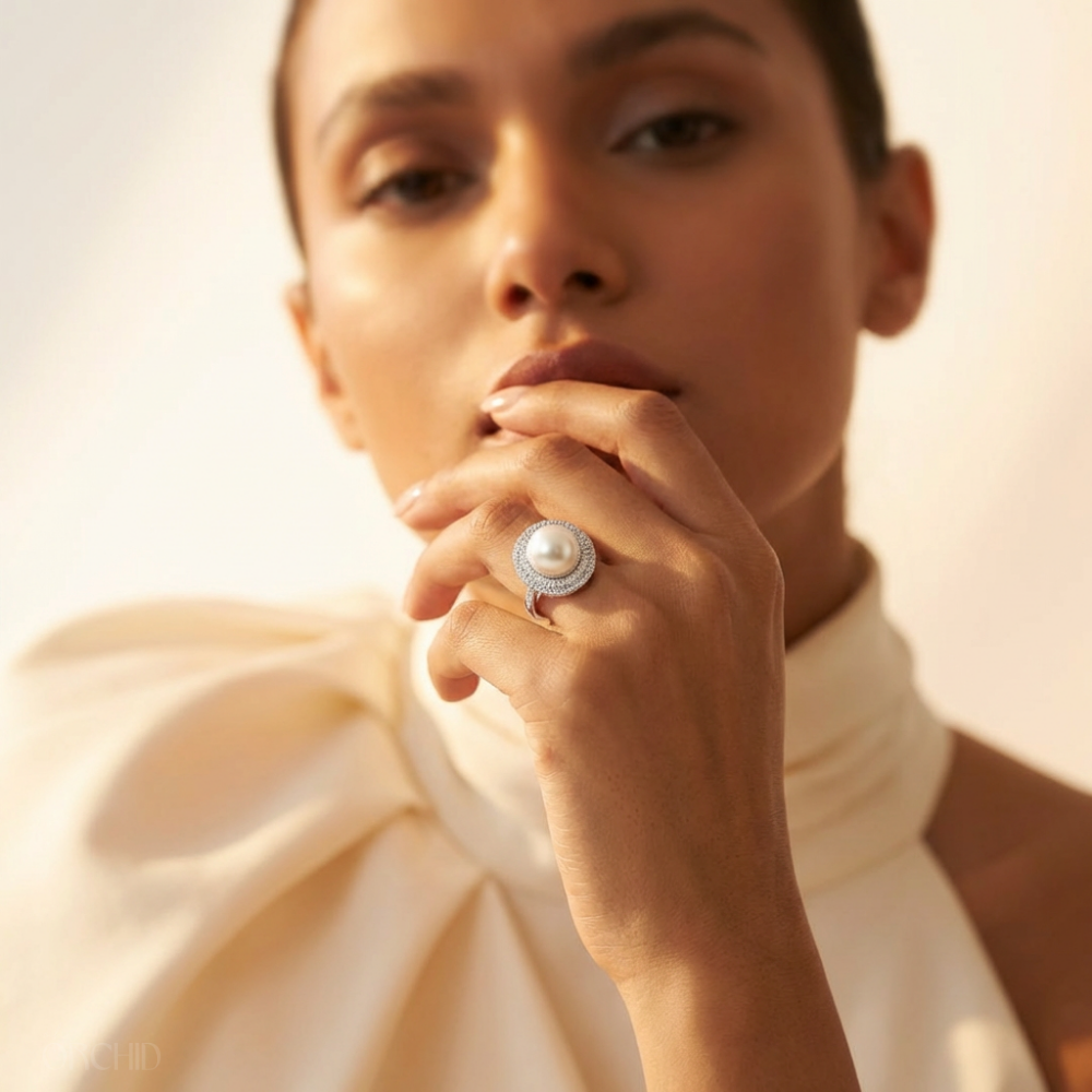 A hand resting against a cheek, wearing a gold ring set with a large pearl