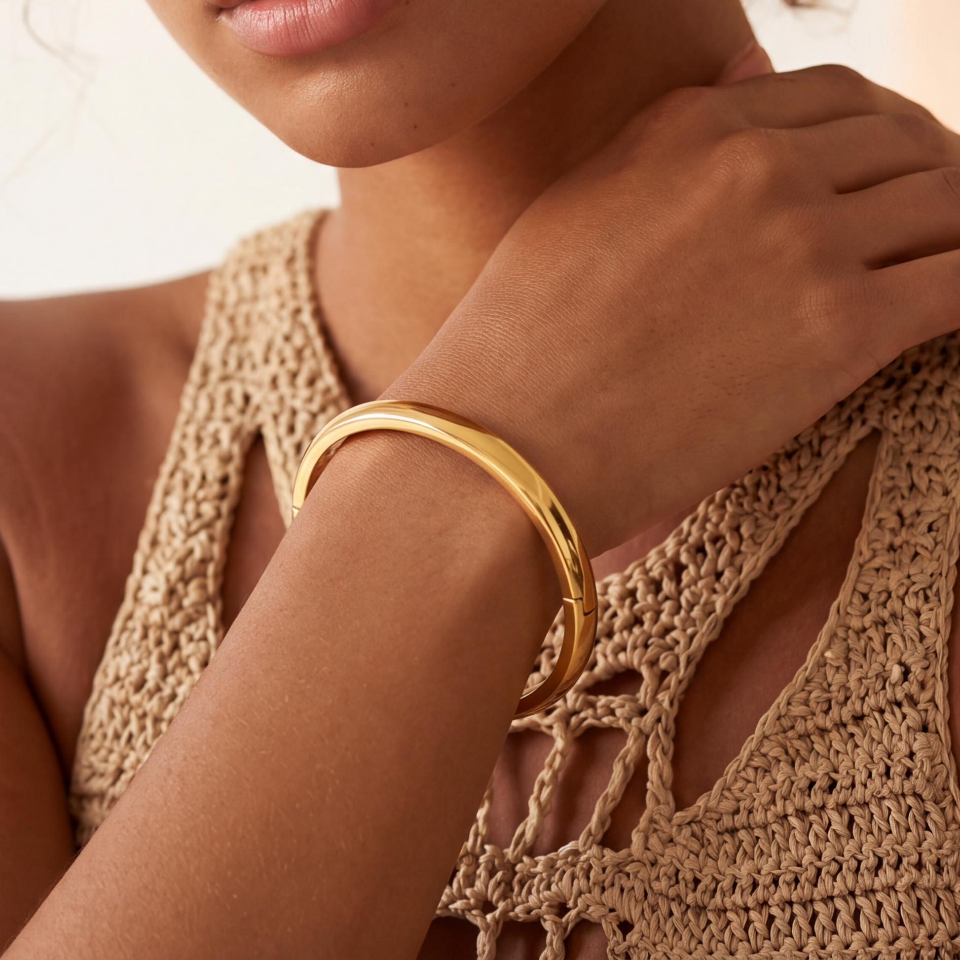 A close-up of a woman wearing a polished gold hinged bangle bracelet on her wrist, paired with a beige crochet halter top.