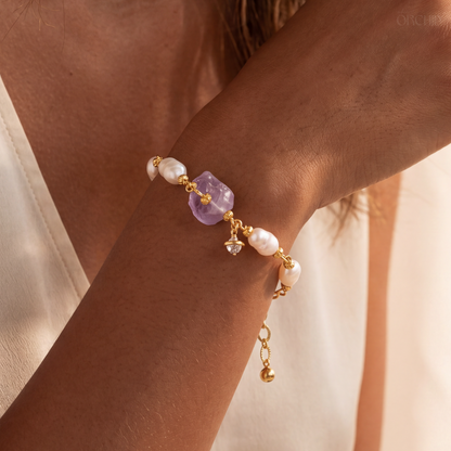 Gold bracelet with pearls and a purple gemstone on a wrist against a neutral background