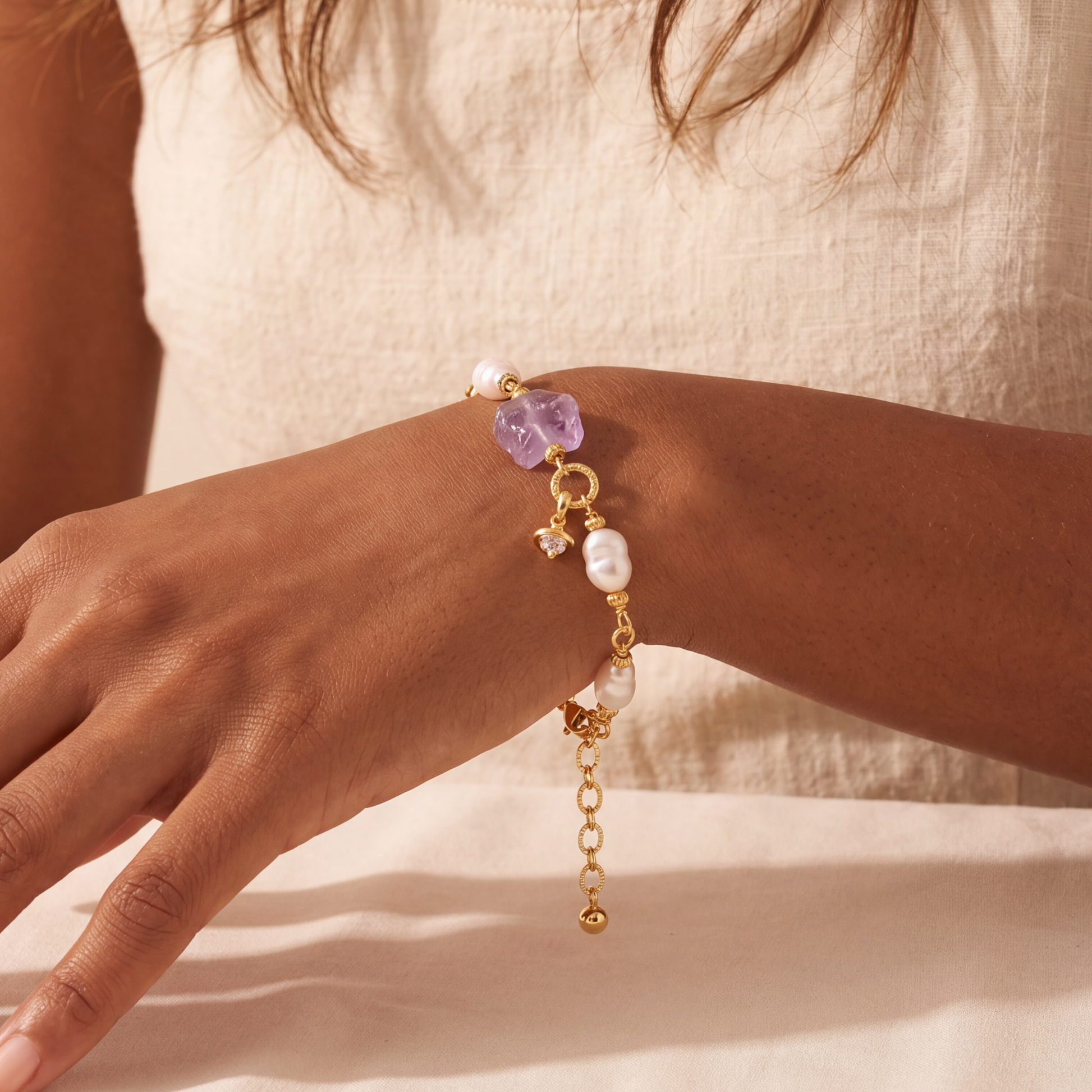 Close-up of a hand wearing a gold bracelet with purple and white beads on a neutral background