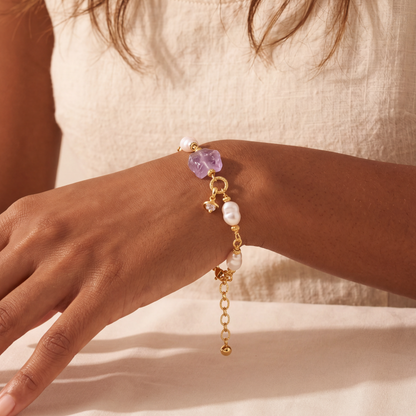 Close-up of a hand wearing a gold bracelet with purple and white beads on a neutral background