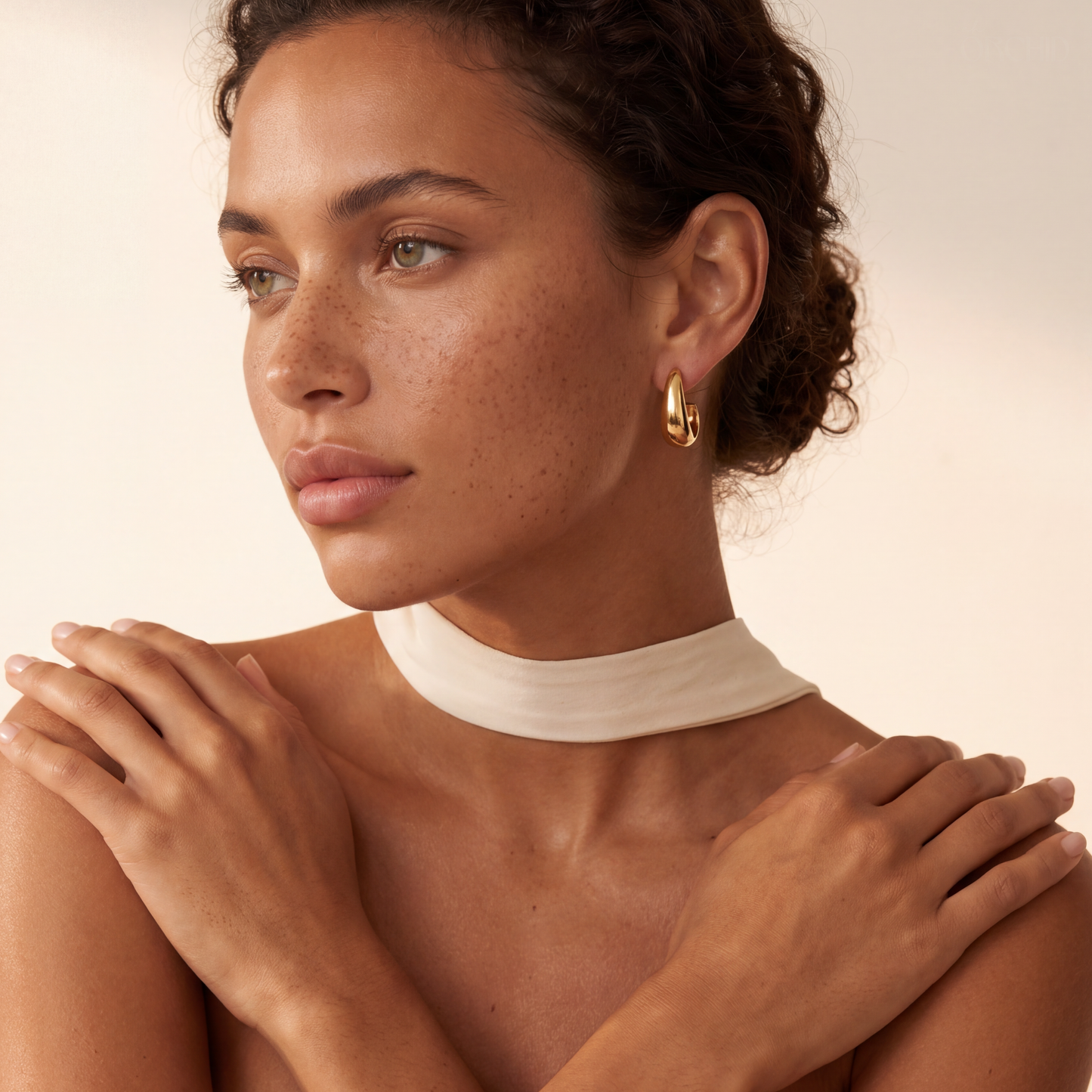 A close-up of a woman wearing a chunky gold hoop earring, with her hands resting on her shoulders and a white high-neck top visible against a warm beige background.