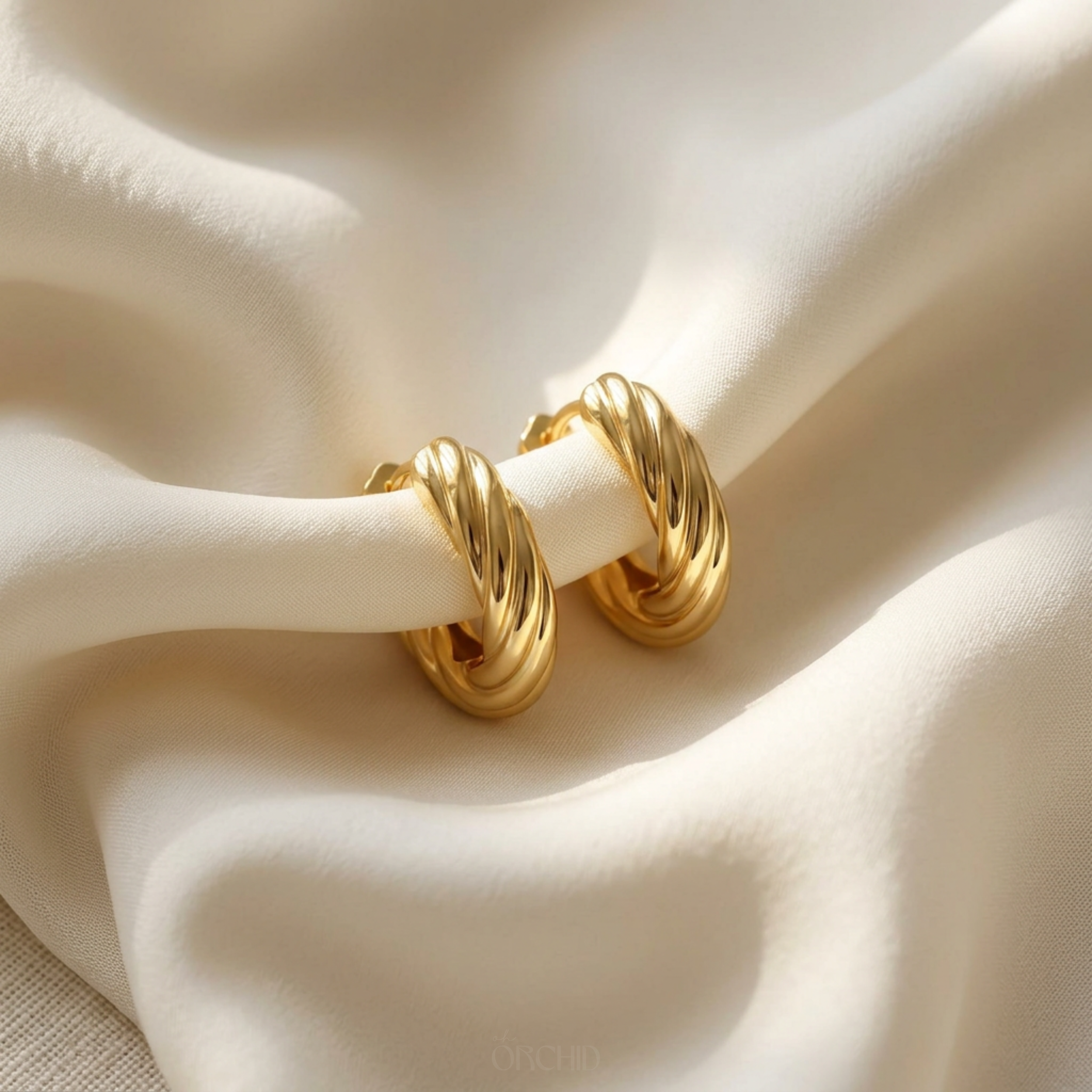 Two twisted gold hoop earrings resting on cream fabric in raking light