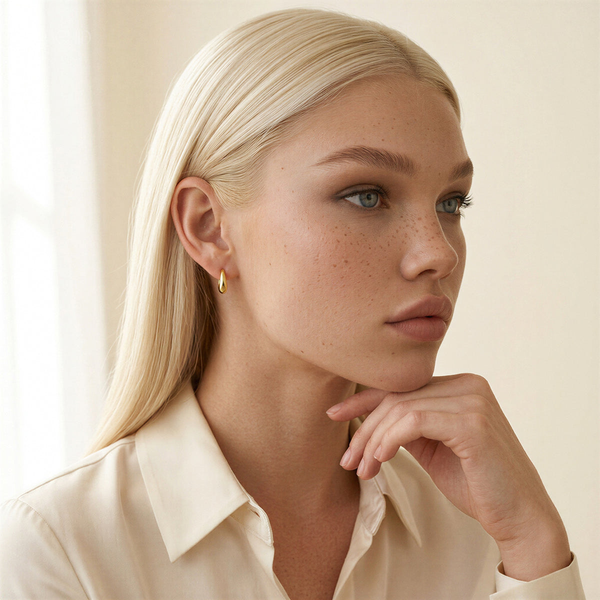 Model in an ivory silk blouse wearing an Aya Huggie Hoop, a small polished 14K gold huggie that sits close to the earlobe
