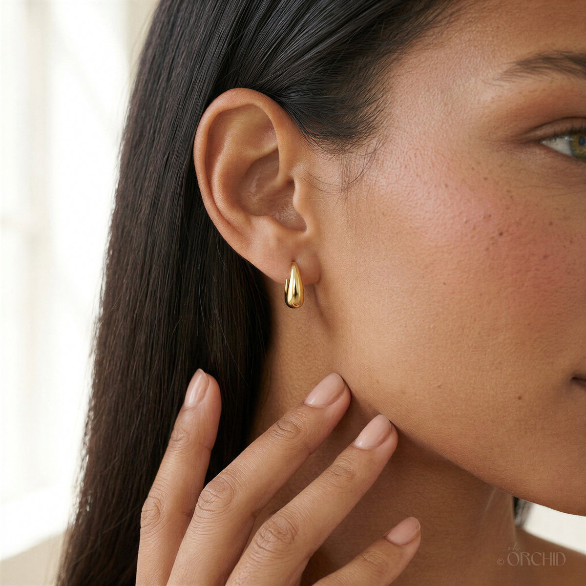 Close-up of an Aya Huggie Hoop on a model's earlobe, a polished 14K gold teardrop huggie about 13 mm long